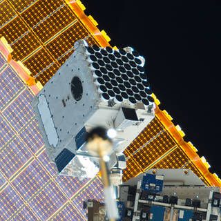 An image of NICER on the exterior of the space station with one of the station’s solar panels in the background